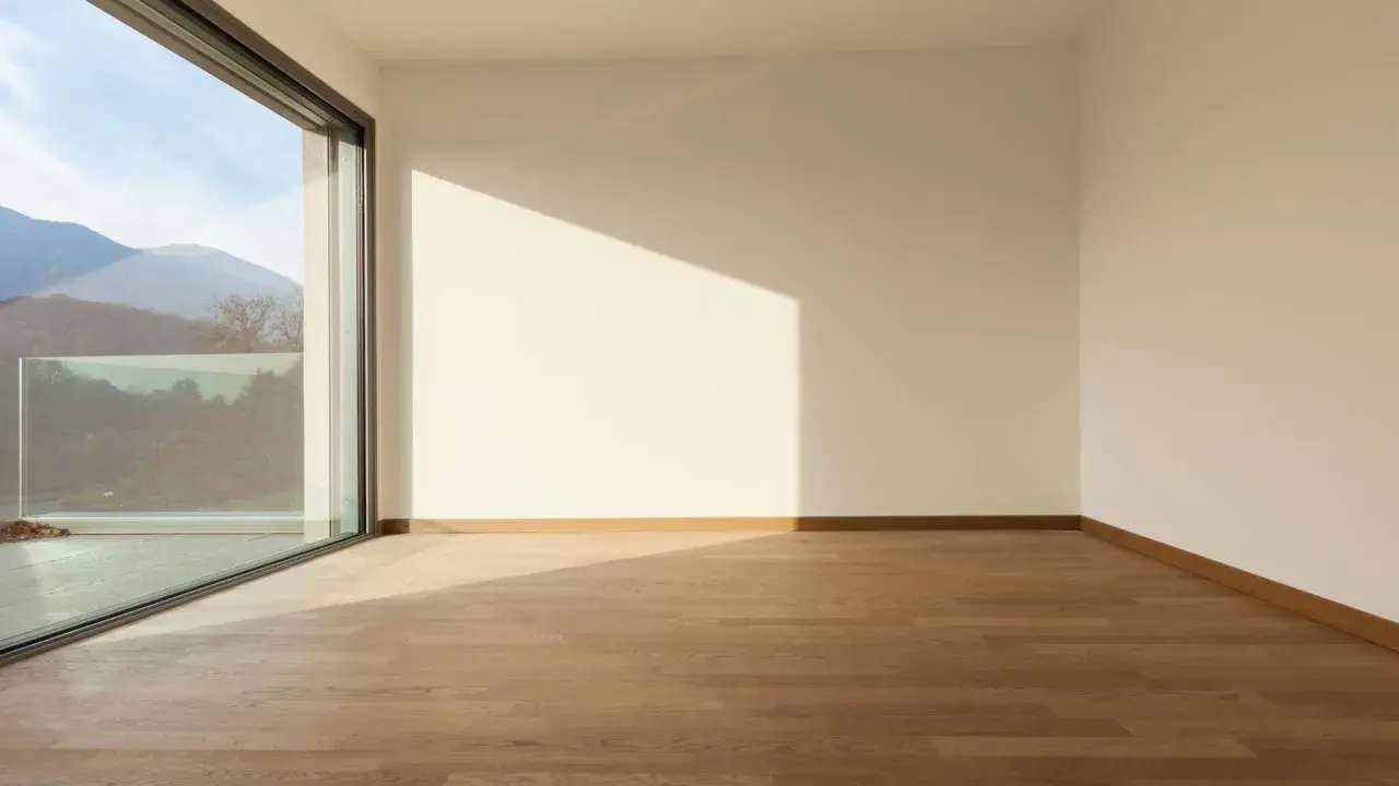 Habitación vacía con suelo de madera y grandes ventanas aislantes que ofrecen vistas a montañas y árboles.