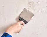 Mano con espátula metálica trabajando en la reparación y alisar paredes con masilla.