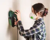 Mujer con mascarilla lijando paredes con una lijadora eléctrica.
