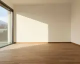 Habitación vacía con suelo de madera y grandes ventanas aislantes que ofrecen vistas a montañas y árboles.