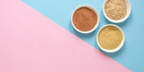 Tres cuencos blancos con polvos de diferentes tonos, como masilla para pared interior, sobre un fondo dividido en rosa y azul.