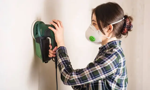 Mujer con mascarilla lijando paredes con una lijadora eléctrica.