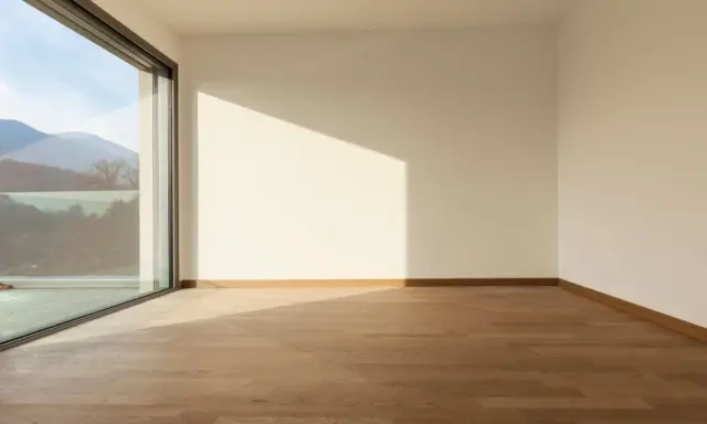 Habitación vacía con suelo de madera y grandes ventanas aislantes que ofrecen vistas a montañas y árboles.