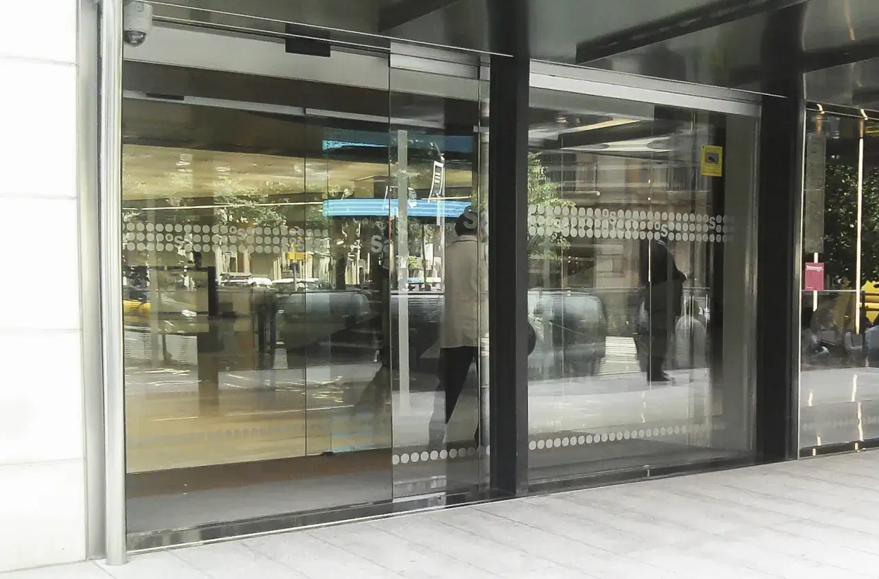 Entrada a un edificio con puertas autom&aacute;ticas de cristal blindado. Reflejos de &aacute;rboles y edificios en el exterior.