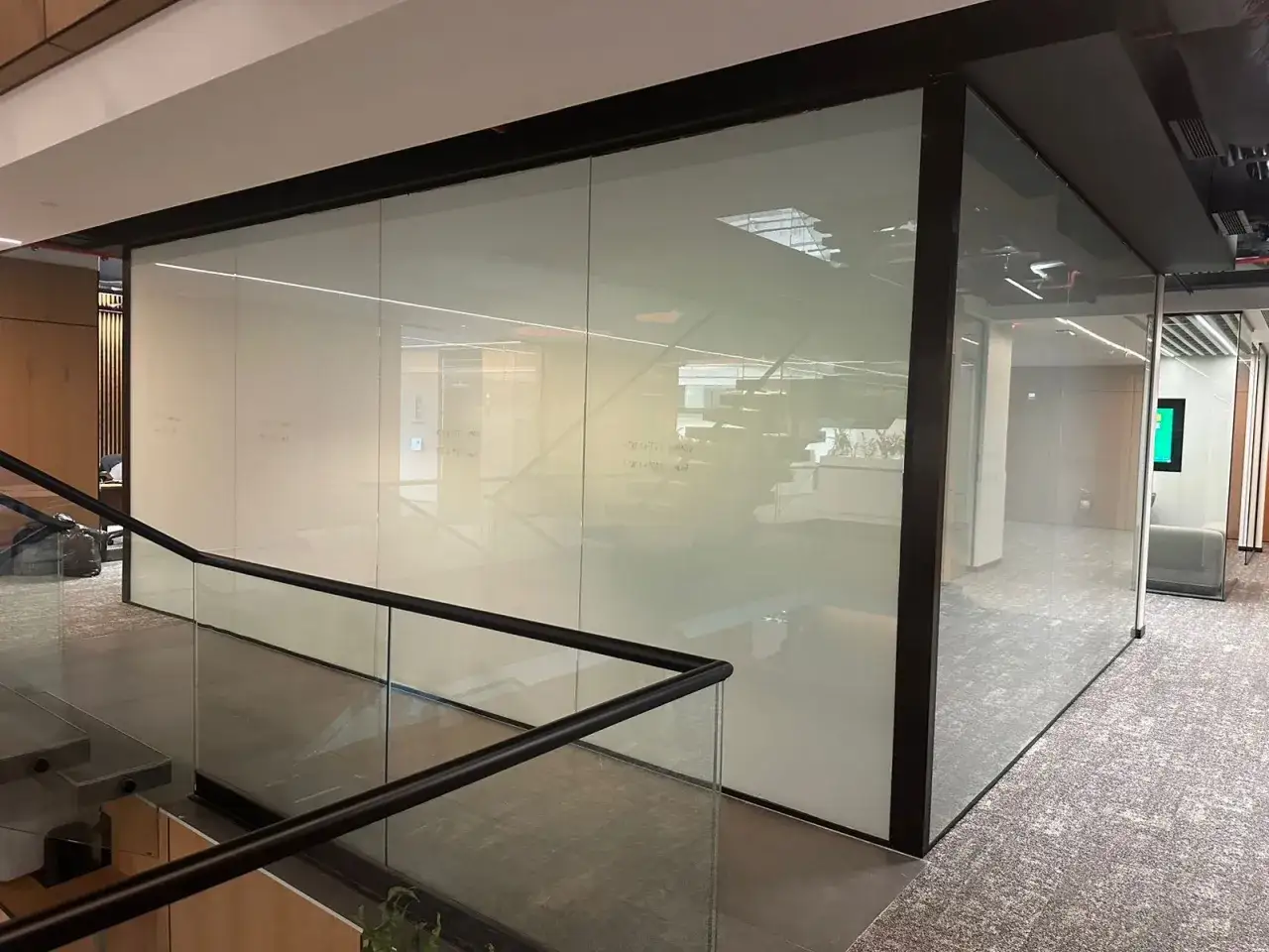 Una oficina moderna con una l&aacute;mina cristal inteligente que divide espacios, reflejando escaleras y mobiliario.