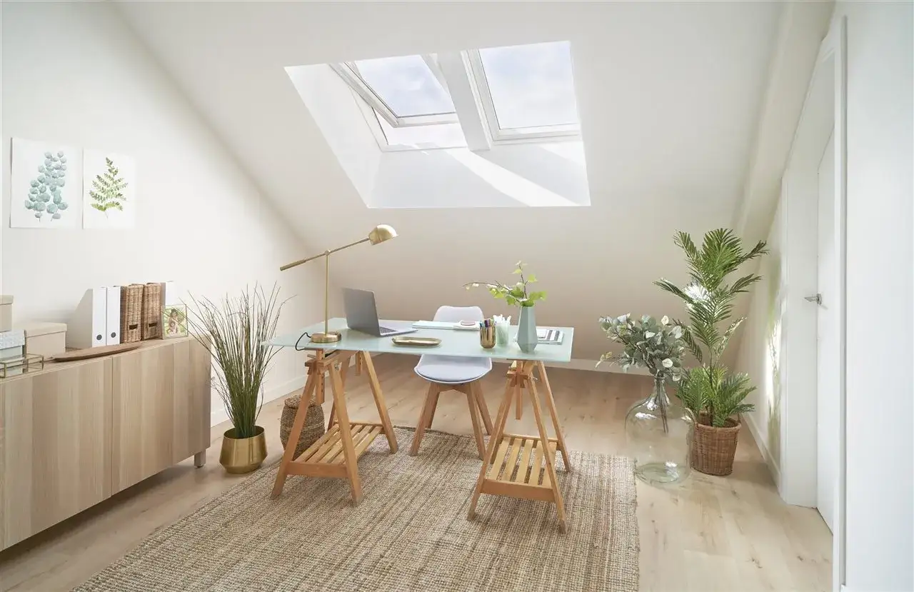 Un estudio luminoso bajo ventanas abuhardilladas, con escritorio, plantas y un mueble de madera.