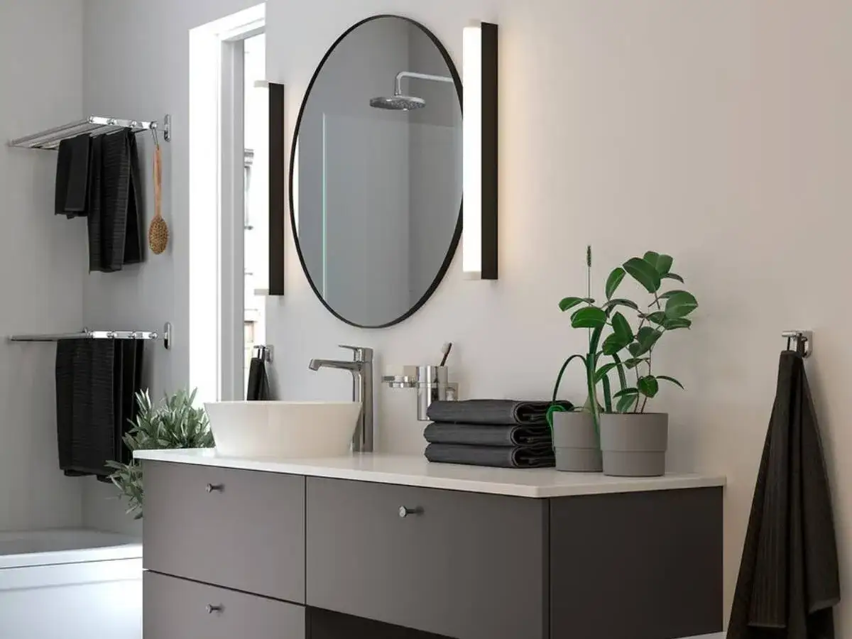 Moderno lavabo con grifería cromada, espejo ovalado y toallas negras. Un toque de verde con plantas. Ideal para una reforma baño pequeño.
