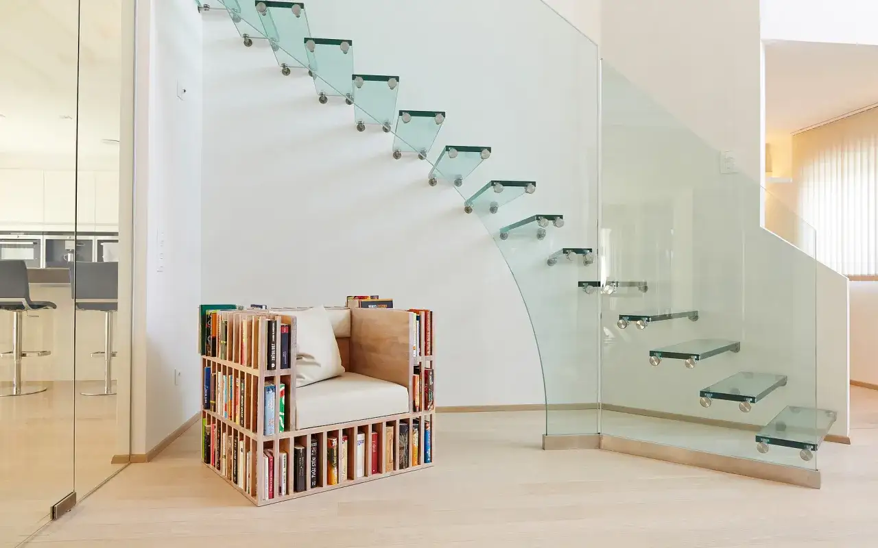 Escalera moderna con pelda&ntilde;os flotantes de cristal y barandillas de cristal. Un sill&oacute;n con estanter&iacute;as integradas para libros se encuentra al pie de la escalera.